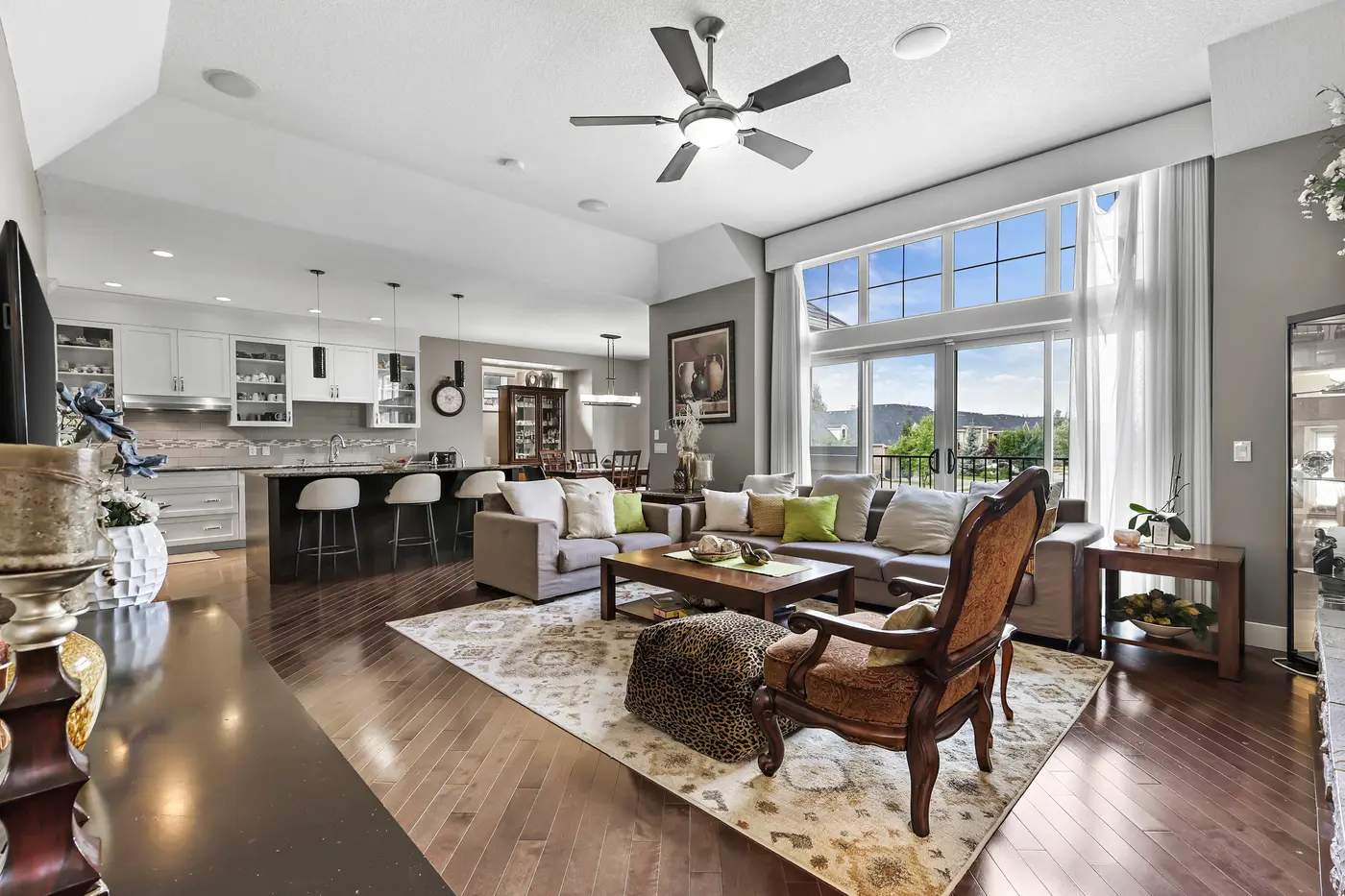 The vaulted great room in full sun — a wall of west-facing windows, sofas gathered on a patterned rug, and the kitchen island and dining area flowing behind.
