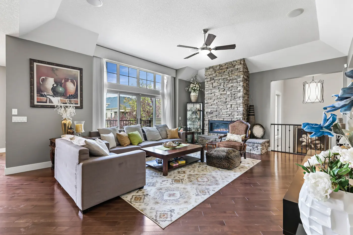 The great room from the fireplace corner — tray ceiling, sectional seating, and the tall windows that hold the afternoon light.