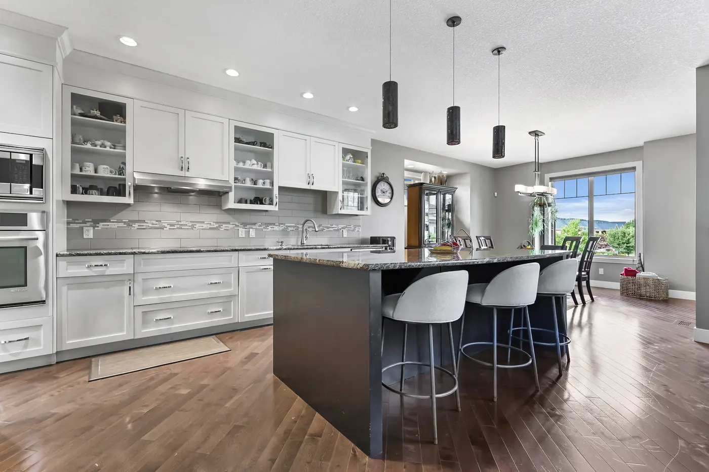 The kitchen — full-height white cabinetry, a long granite-topped island in espresso, cylinder pendants, and seating for three along the bar.