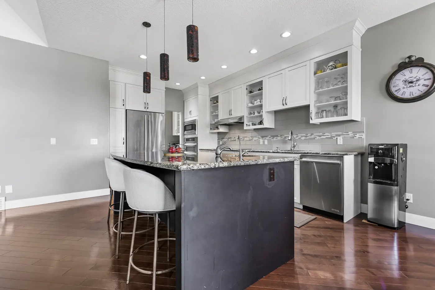 The granite island with upholstered bar stools, glass-front uppers, and the stainless wall ovens and induction cooktop beyond.
