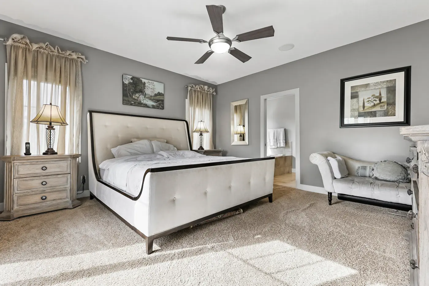 The main-floor primary bedroom — an upholstered wingback bed, a chaise by the window, and the ensuite opening off the far wall.