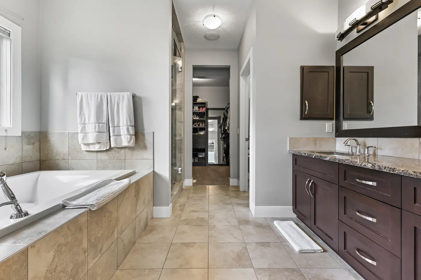 The five-piece ensuite — a tiled soaker tub under the window, a long granite vanity, and the walk-through to the closet beyond.