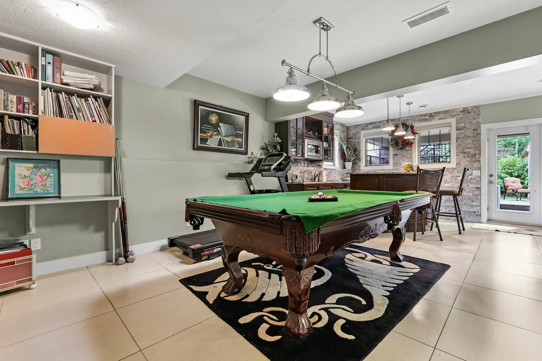 The walkout game room, billiard table at centre, with the stone-clad bar and a glass door to the lower patio beyond.