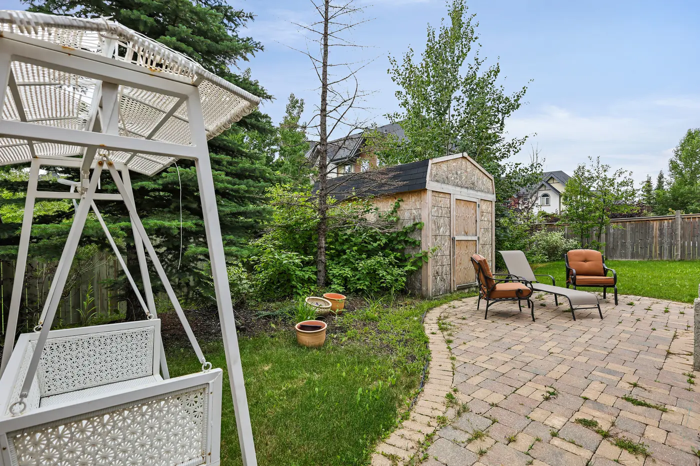 The private backyard — paver path curving past a garden shed, lounge chairs in the sun, and mature trees along the fence.